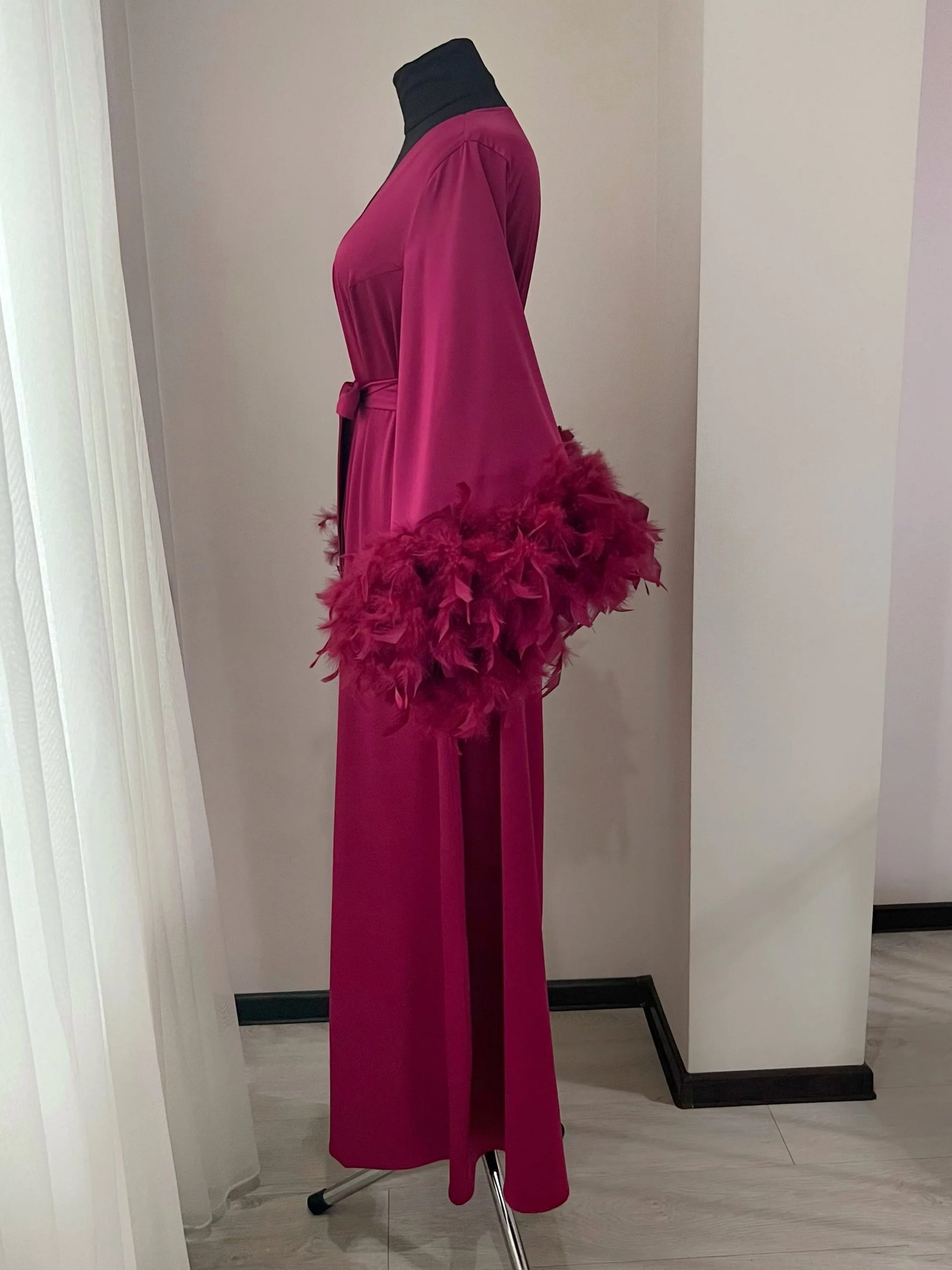 A mannequin wearing a long, burgundy-colored robe with feather accents, displayed in a room with white walls and a white curtain.